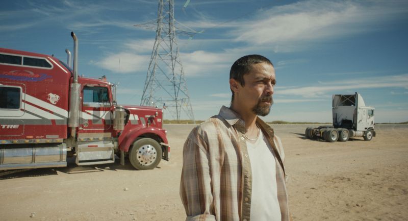 Fotograma oficial de la película "En el camino". Fotograma en plano medio de la película "En el camino". En primer plano a la derecha aparece el actor Víctor Prieto luciendo bigote, barba de candado y vestimenta casual en tonos claros. De fondo, sobre un terreno árido bajo un cielo despejado, se observan un gran tráiler color rojo estacionado a la izquierda, la estructura de una torre de alta tensión al centro y un chasis de camión a la derecha.