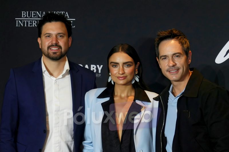 Conferencia de prensa de Hasta el fin del mundo en Mexico Fotografía de tres personas posando sonrientes de medio cuerpo frente a un fondo negro con letras blancas parciales. De izquierda a derecha aparece un hombre con barba vistiendo traje azul y camisa blanca desabotonada, una mujer al centro con blazer claro y aretes grandes, y un hombre a la derecha con chaqueta oscura y playera polo azul claro.