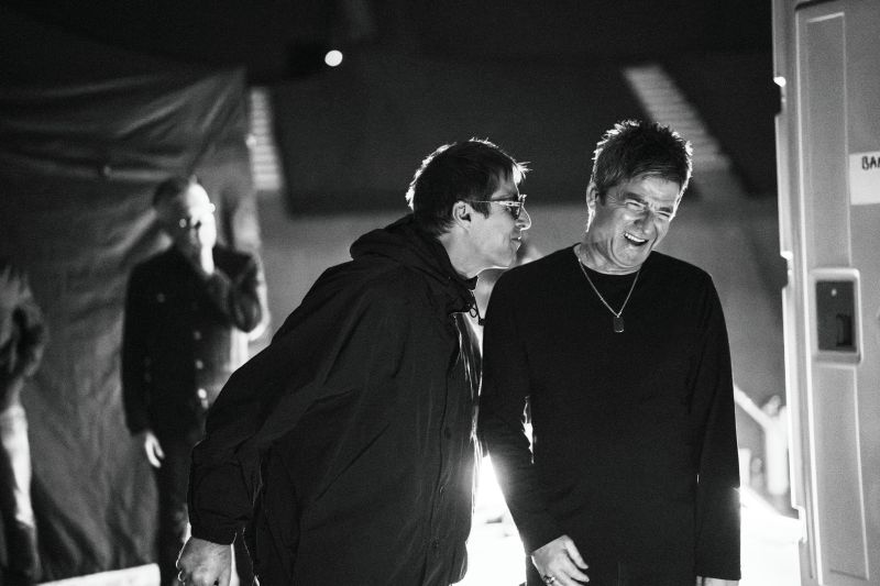 Fotografía en blanco y negro en plano medio durante el backstage de un concierto. Se observa a Liam Gallagher en el centro, vistiendo chamarra de cuello alto y anteojos oscuros, riendo mientras se inclina hacia su hermano Noel Gallagher, quien viste una playera de manga larga, cadena al cuello y sonríe ampliamente. Al fondo a la izquierda se distingue otra figura en la penumbra.