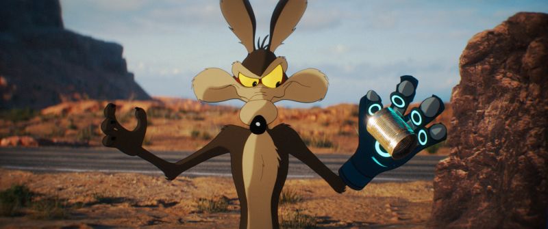 El Coyote en una escena oficial de la pelicula Coyote vs Acme distribuida por Zima Entertainment en Cinepolis Fotografía promocional en plano medio al aire libre con fondo de paisaje desértico de cañones al atardecer. En el centro, el personaje animado del Coyote se muestra parado sobre la carretera con una expresión seria y concentrada mirando de reojo hacia la derecha. Porta un guante tecnológico de color azul con círculos luminosos en su mano izquierda, sosteniendo en la palma una pequeña lata metálica cilíndrica.
