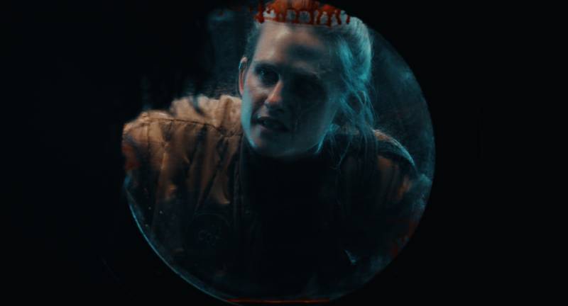 Persona observada a través de una escotilla circular en un ambiente oscuro. Fotografía de un personaje visto a través de una escotilla circular iluminada por un tono azulado, que refuerza la sensación de encierro y tensión en un entorno oscuro.