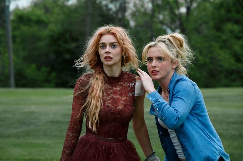 Dos mujeres en un momento de tensión al aire libre Dos mujeres observan con atención algo fuera de cuadro en un entorno natural, una de ellas con vestido rojo y la otra con camisa azul, ambas mostrando preocupación en su expresión.