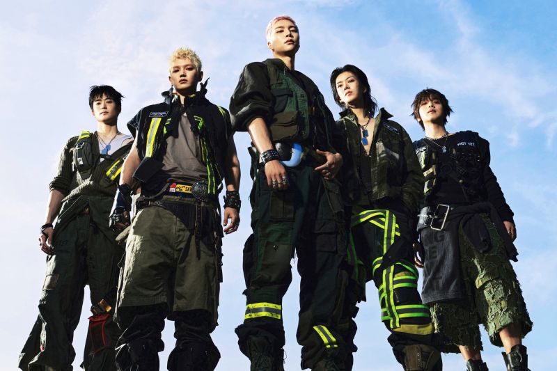 Fotografía promocional de los integrantes de la agrupación de K-pop NCT 127. Aparecen cinco miembros posando en contrapicado bajo un cielo azul despejado, luciendo atuendos de estilo industrial y táctico en tonos verde oliva, negro y beige con cintas fluorescentes.