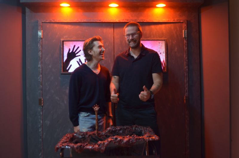 El actor Austin Abrams y el director Zach Cregger posan sonrientes y de pie en un set temático decorado con iluminación roja y elementos de utilería de terror durante la presentación de la película en Ciudad de México.