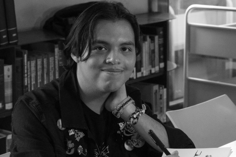 Fotografía en blanco y negro del artista y animador poblano Aarón Cruz sonriendo a la cámara mientras sostiene un bolígrafo sobre su libreta de bocetos. Viste una chamarra con pines y accesorios en la muñeca, sentado en una mesa con libros al fondo.