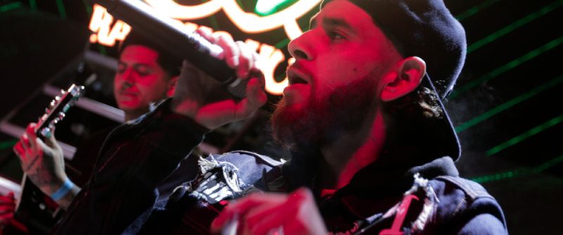 Actuación en vivo – Clika Cantante en escenario con iluminación roja y verde acompañado de guitarrista.