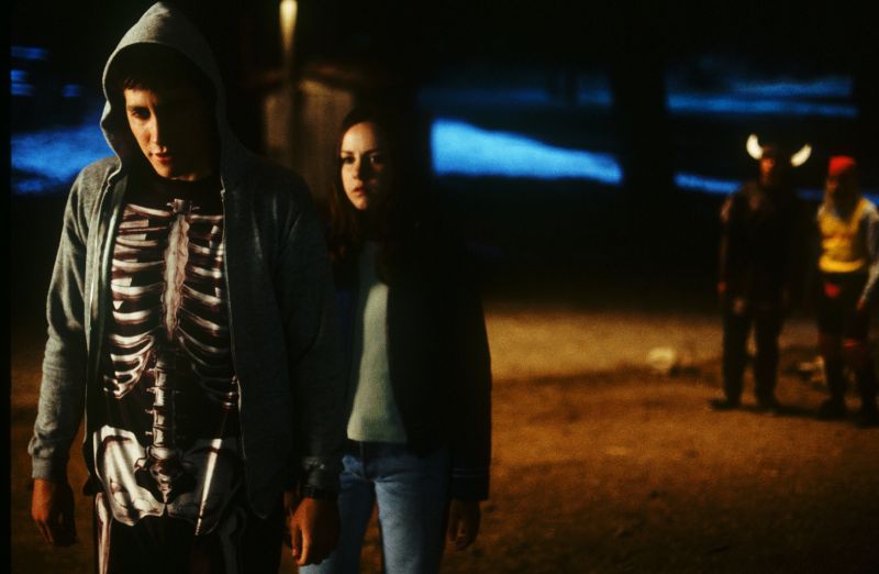 Escena nocturna – Donnie Darko Grupo de jóvenes al aire libre durante la noche, con disfraces y vestimenta casual en un ambiente oscuro y ligeramente inquietante.