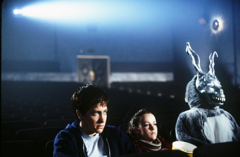 Frank en la sala de cine – Donnie Darko Tres personas en una sala de cine vacía, una de ellas usando un traje de conejo metálico que genera un ambiente surreal.