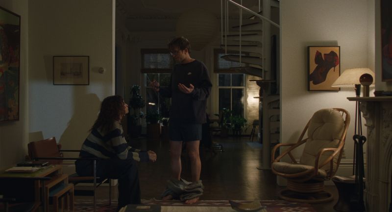 Encuentro en sala doméstica – El Drama Dos personas en una sala cálida: una sentada con suéter a rayas y otra de pie frente a ella, con pantalones a la altura de los tobillos. El entorno muestra muebles de madera, una escalera de caracol y decoración hogareña.