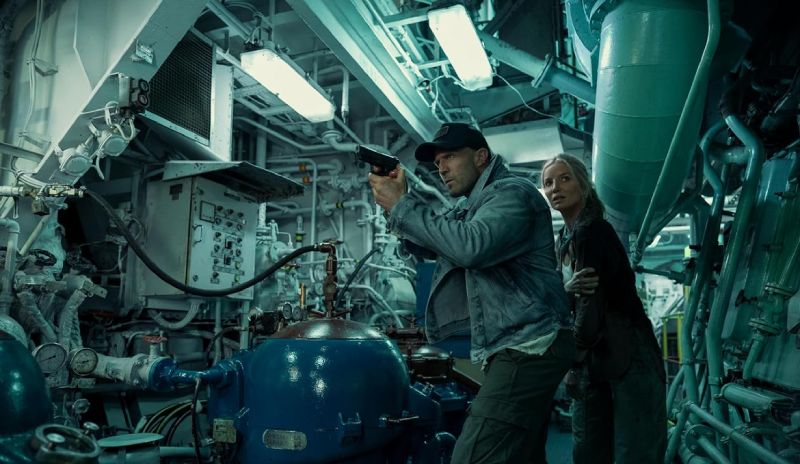 Jason Statham apuntando con una pistola dentro de la sala de máquinas de un barco mientras protege a una mujer detrás de él.