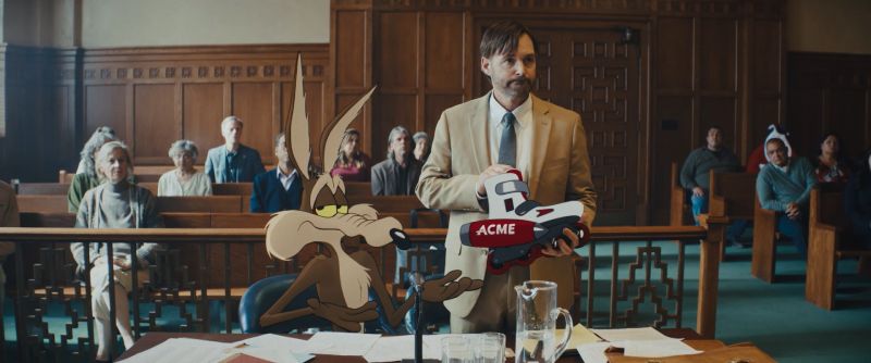 El Coyote y su abogado en el tribunal durante el juicio contra ACME Will Forte sosteniendo un patín defectuoso marca ACME en un tribunal junto al Coyote animado, quien señala resignado ante la sala.