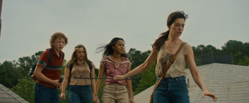 Grupo de personajes sobre el tejado en El final de la Calle Oak Fotograma de Anne Hathaway al frente con una vara de metal ensangrentada guiando a tres jóvenes sobre el tejado de una vivienda mientras observan alerta su entorno.