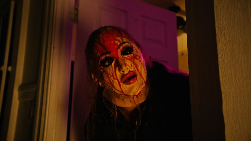 Fotograma en plano medio en penumbra con luz morada y cálida, donde un personaje asoma la cabeza por el marco de una puerta vistiendo una máscara femenina manchada con abundante sangre roja en la frente.