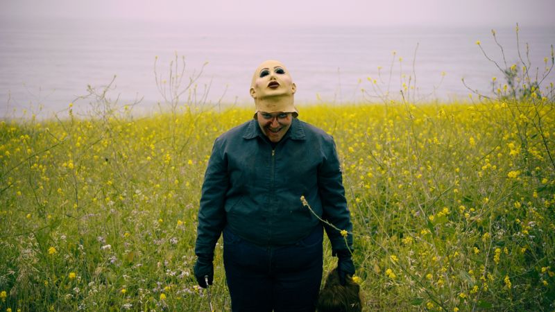 Plano general de un personaje en medio de un campo de flores amarillas bajo un cielo nublado, vistiendo una chamarra oscura y una máscara de manicura pálida levantada hacia la frente, revelando una sonrisa desquiciada y sangrienta.