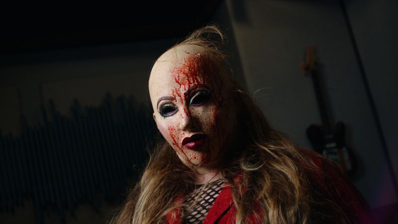 Primer plano en un estudio de grabación con guitarras al fondo, mostrando a la protagonista con peluca rubia y una máscara con salpicaduras de sangre cayéndole por el rostro.