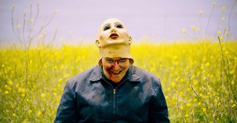 Plano medio corto en un campo abierto donde la persona sonríe con la boca manchada de sangre mientras mantiene la máscara de látex levantada sobre la cabeza a la altura de la frente.
