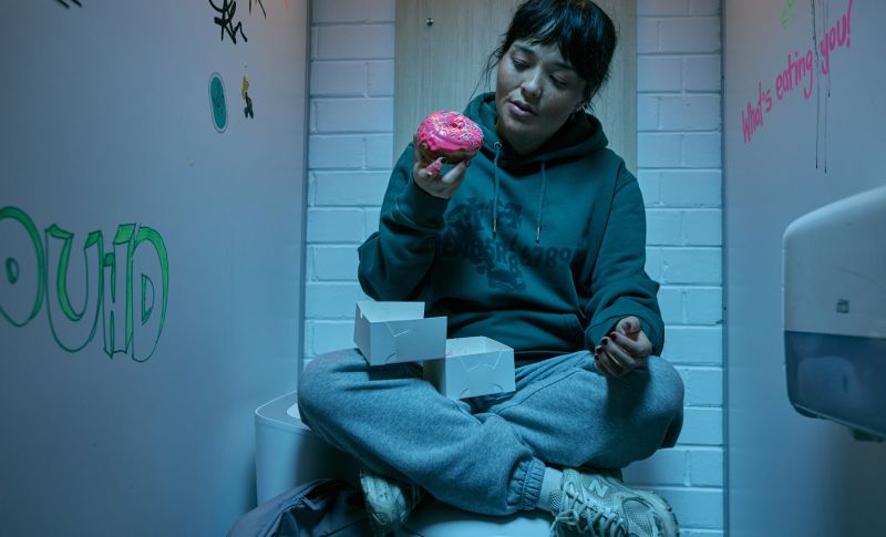 Hanna comiendo a escondidas en el baño Hanna (Midori Francis) sentada con las piernas cruzadas sobre un inodoro en el cubículo de un baño, sosteniendo una dona rosada con chispas extraída de una caja de repostería.