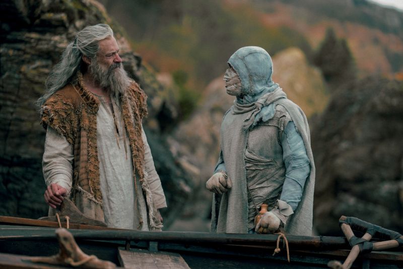 Robin Hood y un misterioso personaje en La Muerte de Robin Hood Robin Hood (Hugh Jackman) de pie junto a un bote frente a un hombre, quien viste ropas raídas y vendajes grises cubriéndole la cabeza y el cuerpo.