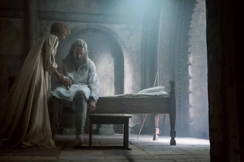 Jodie Comer atiende a Hugh Jackman en La Muerte de Robin Hood Interior de una estancia medieval donde una mujer vestida con una túnica clara atiende las heridas del brazo de Robin Hood, quien está sentado exhausto en la cama.