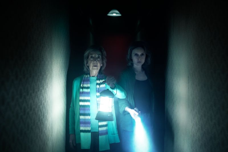 Elise Rainier acompañada de Gemma recorriendo un pasillo oscuro iluminado únicamente por una linterna de gas y una linterna de mano.