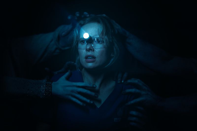 Gemma con una lámpara frontal de dentista en la cabeza mientras múltiples manos espectrales y oscuras emergen de las sombras para sujetarla.