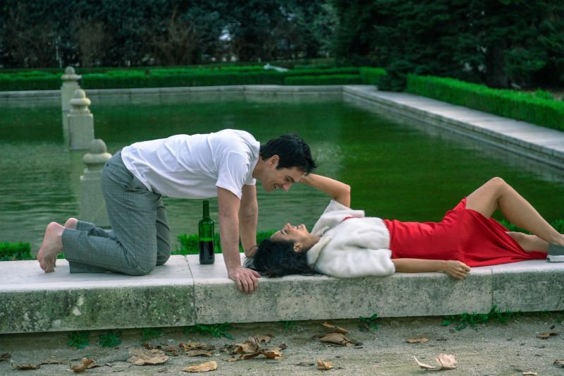 Fotograma de Hasta el fin del mundo donde Aislinn Derbez yace acostada con un vestido rojo sobre el borde de una fuente de piedra mientras Mauricio Ochmann se inclina sonriente hacia ella.