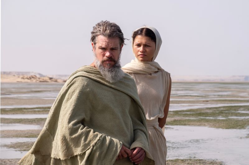 Matt Damon como Odiseo y Zendaya como Atenea en La Odisea Fotograma de la película La Odisea donde se observa a Matt Damon con vestimenta clásica de la Grecia antigua y barba canosa representando a Odiseo, acompañado por Zendaya con túnica y velo blanco en una locación natural costera.