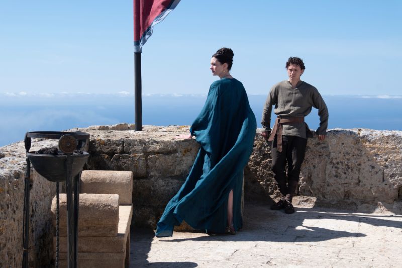 Anne Hathaway como Penélope y Tom Holland como Telémaco en La Odisea Escena del filme La Odisea con Anne Hathaway luciendo un vestido azul estilo griego y Tom Holland como su hijo Telémaco en la terraza de una fortaleza de piedra junto al mar.