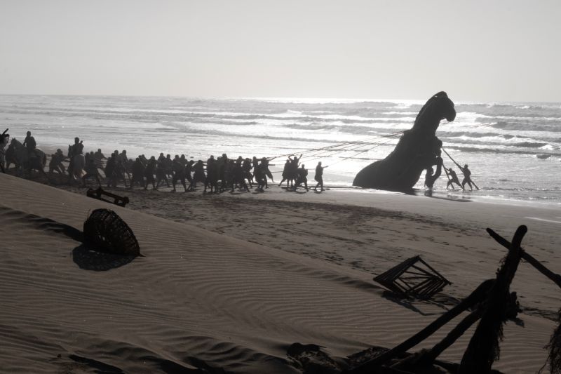 Secuencia del Caballo de Troya en La Odisea de Christopher Nolan Captura cinematográfica en blanco y negro que muestra a decenas de hombres arrastrando la estructura del Caballo de Troya a lo largo de una playa durante una de las secuencias de flashback de la película.