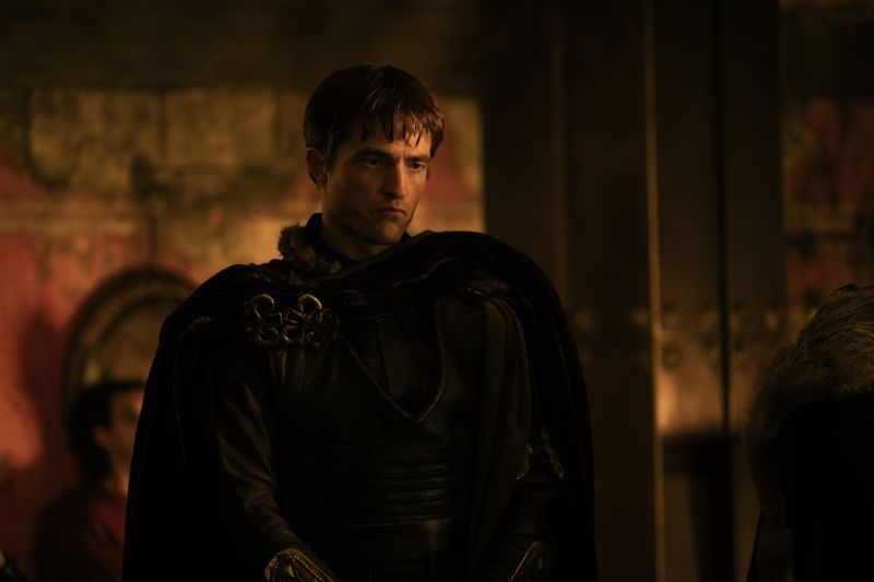 Robert Pattinson interpreta a Antínoo en La Odisea Primer plano medio de Robert Pattinson caracterizado como Antínoo en una escena nocturna/interior luciendo vestimenta de cuero y capas obscuras en la película La Odisea.