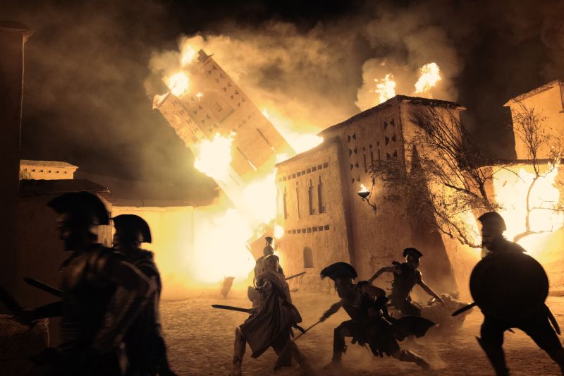 Secuencia de batalla e incendio en La Odisea de Nolan Plano de acción de La Odisea que capta a varios soldados con cascos griegos combatiendo en medio de un incendio nocturno con una torre cayendo en llamas tras ellos.