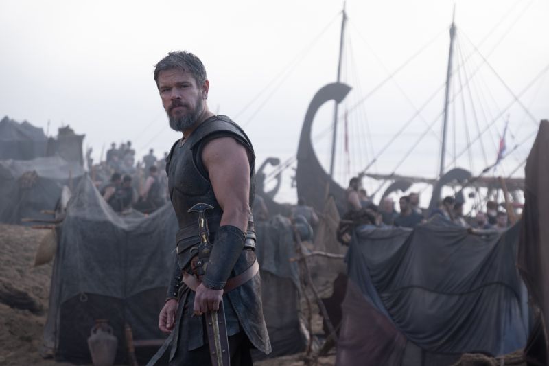 Matt Damon como Odiseo en el campamento militar griego Fotograma de la película La Odisea donde aparece Matt Damon en primer plano portando armadura de combate antigua y sosteniendo una espada, situado en una playa repleta de tiendas de campaña y trirremes griegos.