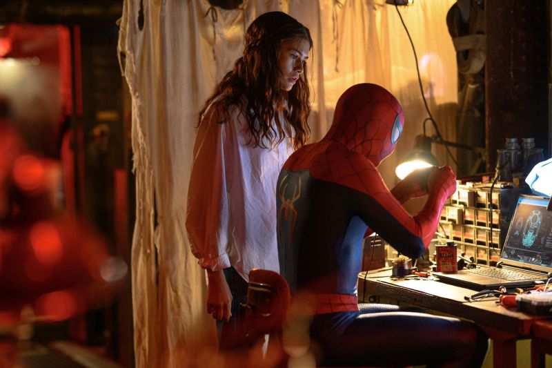 Peter Parker y MJ en el taller de trabajo Peter Parker usando el traje de Spider-Man sentado en una mesa de trabajo reparando equipo tecnológico frente a una laptop, mientras MJ (Zendaya) lo observa atentamente.