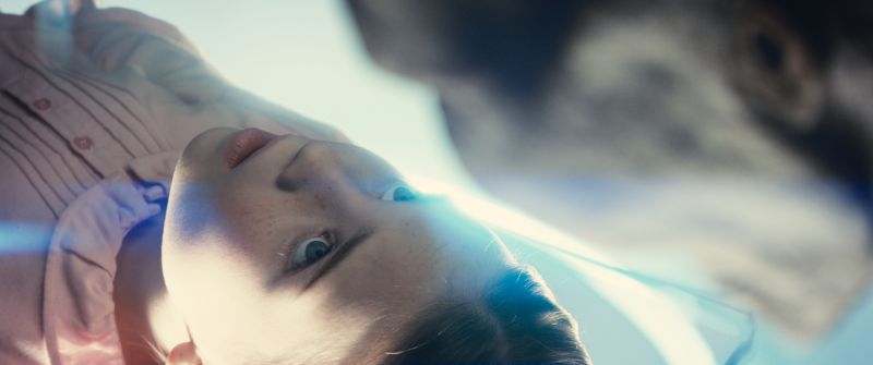 Conexión íntima entre personaje y extraterrestre Joven acostado observando de cerca a un extraterrestre en un entorno luminoso.