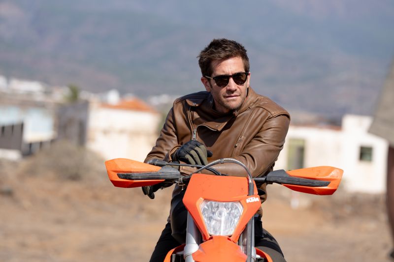 Persona con chamarra de cuero y lentes oscuros sobre una motocicleta en un paisaje árido.