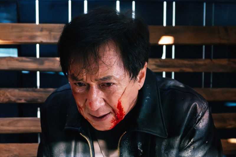Wong Tak-chung bajo presión — Enemigo Mortal Hombre con chaqueta de cuero y rostro ensangrentado dentro de una estructura de madera iluminada dramáticamente.