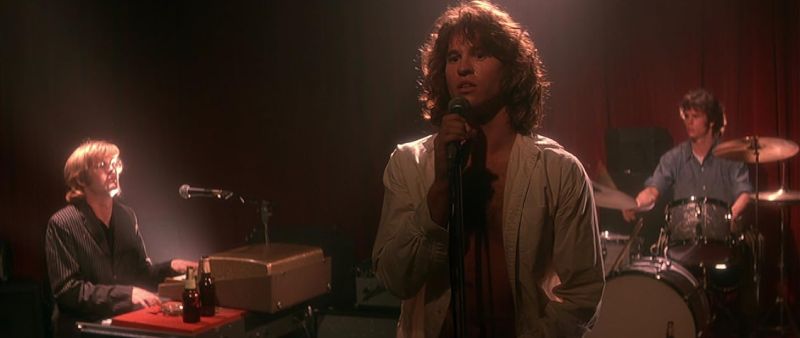 Escena musical con cantante al centro, tecladista a la izquierda y baterista al fondo sobre un escenario iluminado.