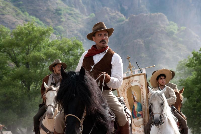 Andy García como el General Gorostieta Andy García caracterizado como el General Enrique Gorostieta montando a caballo en el campo junto a tropas cristeras y un estandarte religioso.