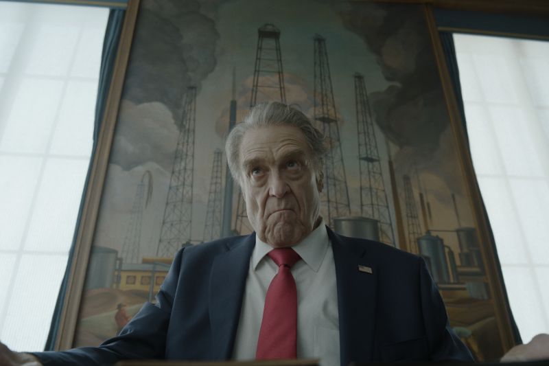 John Goodman como el Presidente Compson en Digger John Goodman como el Presidente Compson mirando fijamente hacia abajo con expresión seria frente a una pintura de torres de extracción petrolera.