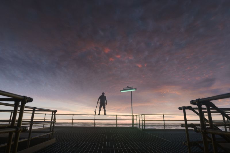 Fotografía de Emmanuel Lubezki para Digger Silueta de un personaje sosteniendo una pala sobre una plataforma industrial en el mar al atardecer bajo un cielo en tonos violeta y naranja.