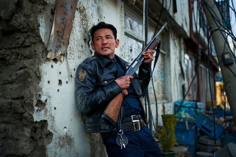 Hwang Jung-min en HOPE: El Primer Impacto El actor Hwang Jung-min vistiendo chamarra de piel y uniforme cubriéndose contra un muro destruido sosteniendo una escopeta.