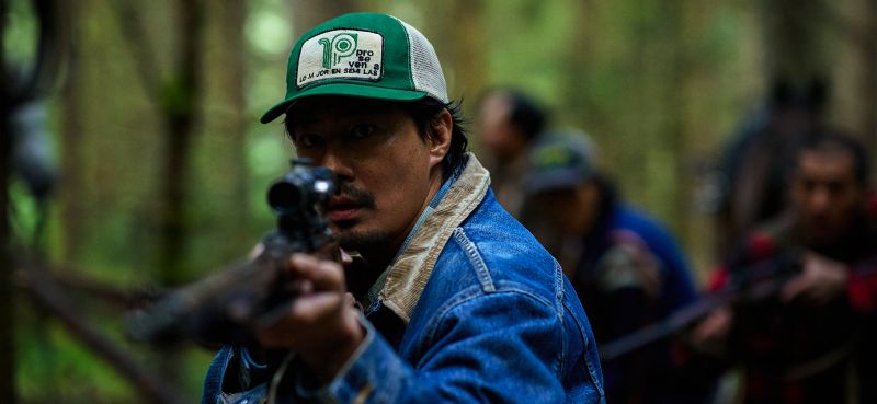 Zo In-sung en HOPE: El Primer Impacto El actor Zo In-sung usando una gorra verde y chamarra de mezclilla apuntando fijamente con un rifle de francotirador en el bosque.
