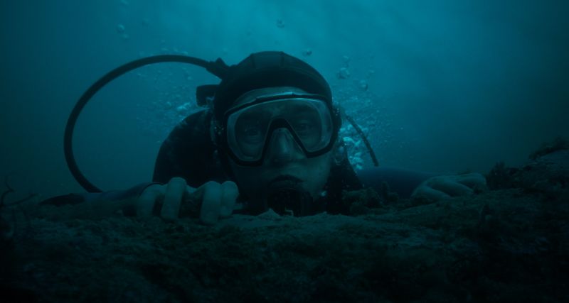 Austin Abrams con equipo completo de buceo mirando con expresión de temor a través del visor bajo el agua.