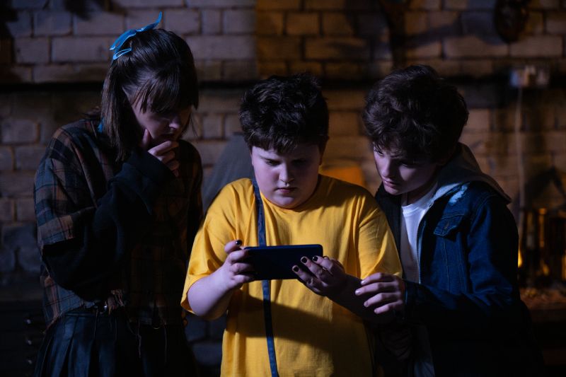 James y sus amigos en una escena de misterio Tres niños reunidos en un sótano iluminados por la luz de una pantalla de teléfono celular que sostienen en el centro.