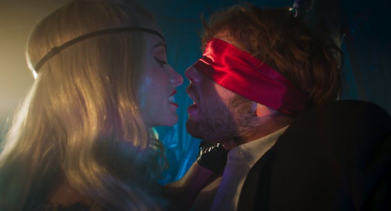 Seducción y Tensión en la Trama Una mujer rubia con venda roja en los ojos e inclinada hacia adelante compartiendo un momento de tensión y cercanía con un hombre.
