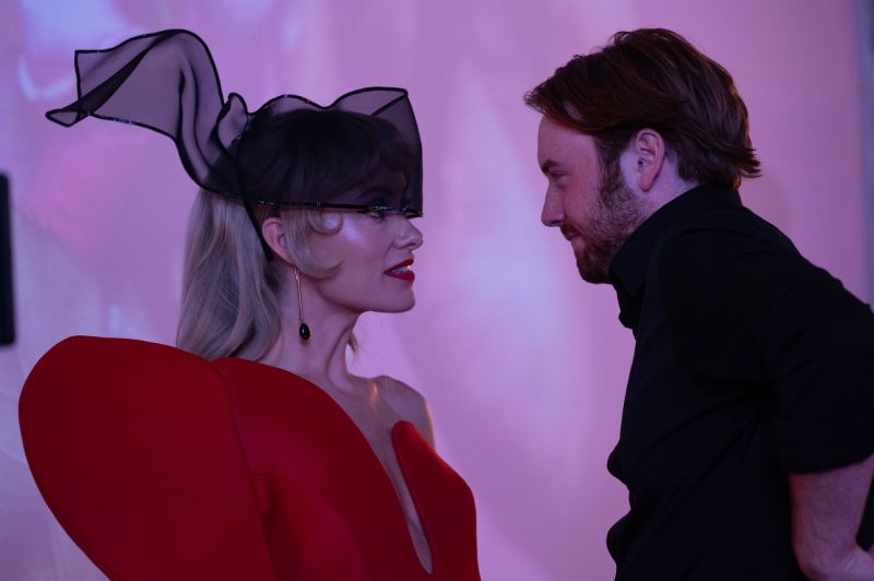 Olivia Wilde y Cooper Hoffman en Escena Olivia Wilde luciendo un vestido rojo con un tocado negro de malla junto a Cooper Hoffman en una habitación iluminada en tonos rosados.