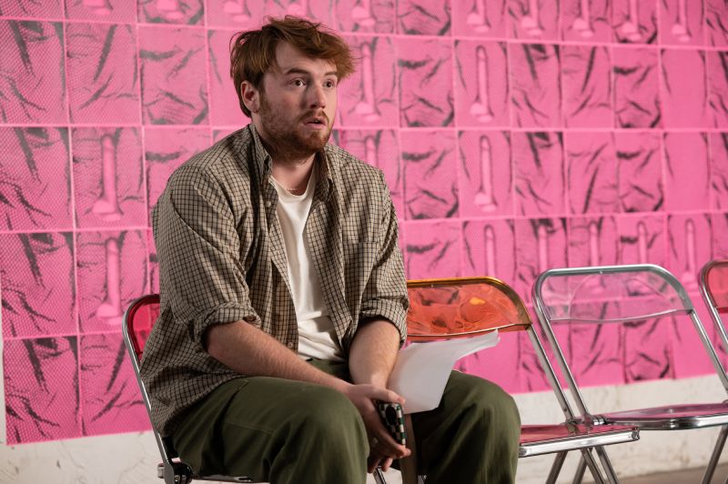 Momento de Incomodidad para el Protagonista Cooper Hoffman sentado con expresión de desconcierto frente a una pared decorada con patrones rosados.