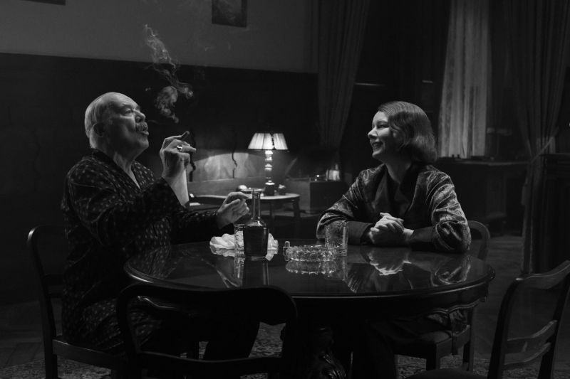 Instantes de Intimidad y Calma Thomas Mann fumando un puro y conversando sonriente con una mujer en una habitación iluminada tenuemente por una lámpara de mesa.