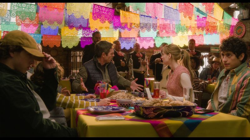 Comida Familiar con Mariachi Escena de discusión en una mesa de restaurante decorada con papel picado y un mariachi tocando al fondo.
