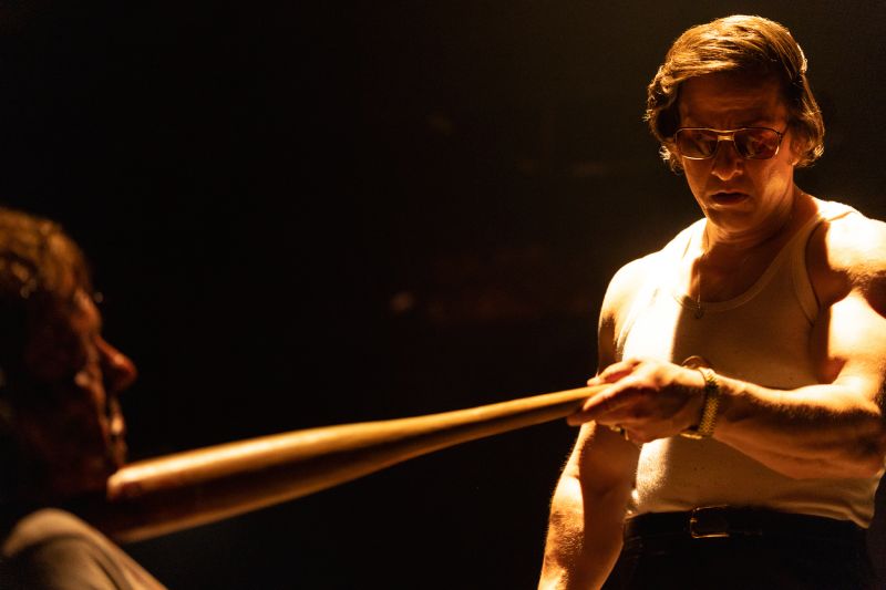 Mark Wahlberg en una habitación oscura usando una camiseta sin mangas y lentes oscuros mientras apunta directamente un bate de béisbol de madera a la garganta de un hombre.